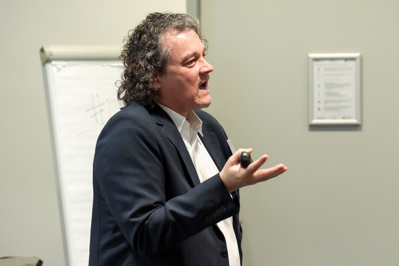 Person mit lockigem Haar und dunklem Sakko gestikuliert vor einem Flipchart in einem Raum mit grauer Wand.