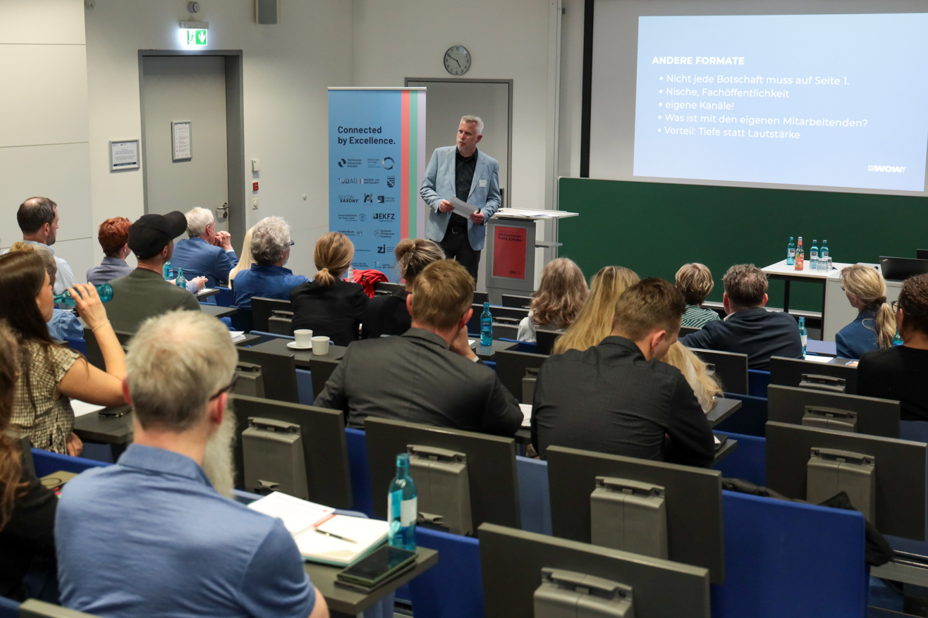 Vortragender steht vor einer grünen Tafel und spricht zu einem Publikum in einem Seminarraum.