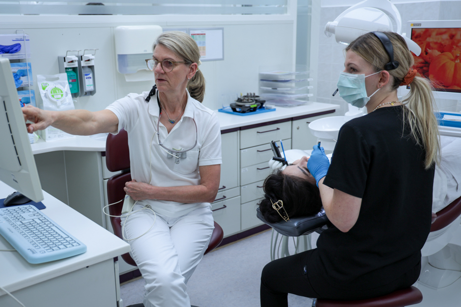 Eine Zahnärztin erklärt etwas am Computer, während eine Kollegin eine Behandlung bei einer Patientin durchführt.