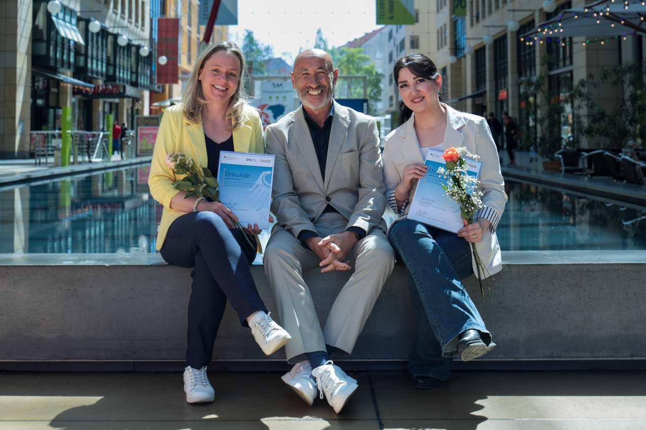 Drei Personen sitzen auf einer Bank in einer Fußgängerzone, zwei halten Blumen und Urkunden.