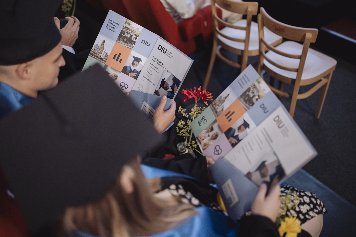 Zwei Absolventen in Talaren halten Urkundenmappen mit dem Schriftzug 'DIU' und sitzen nebeneinander in einem Raum mit Holzstühlen.