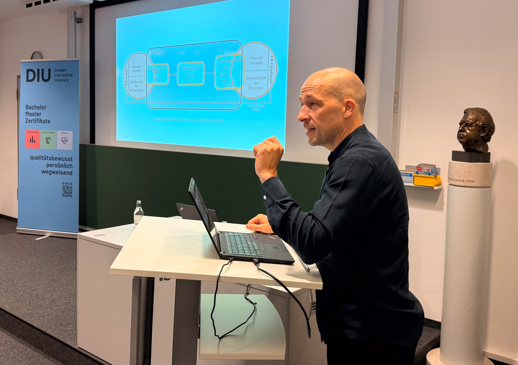 Mann mit schwarzem Hemd hält Vortrag an Stehpult mit Laptop vor Präsentationsbildschirm.