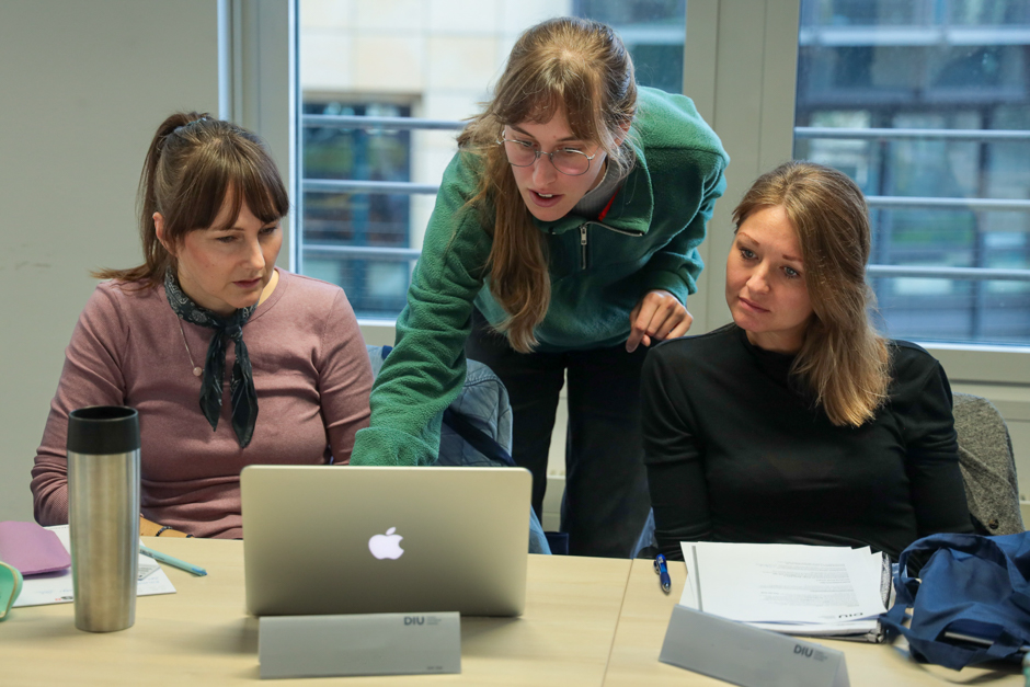 Drei Frauen sitzen an einem Tisch und schauen gemeinsam auf einen Laptop, eine Frau zeigt auf den Bildschirm