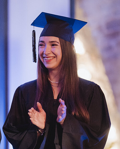 Young woman in graduation gown and cap clapping and smiling indoors