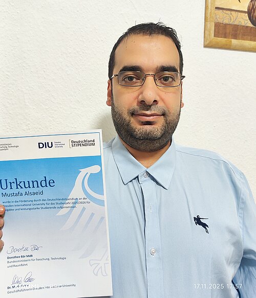 Man wearing glasses and a light blue polo shirt holding a certificate from DIU Deutschland Stipendium