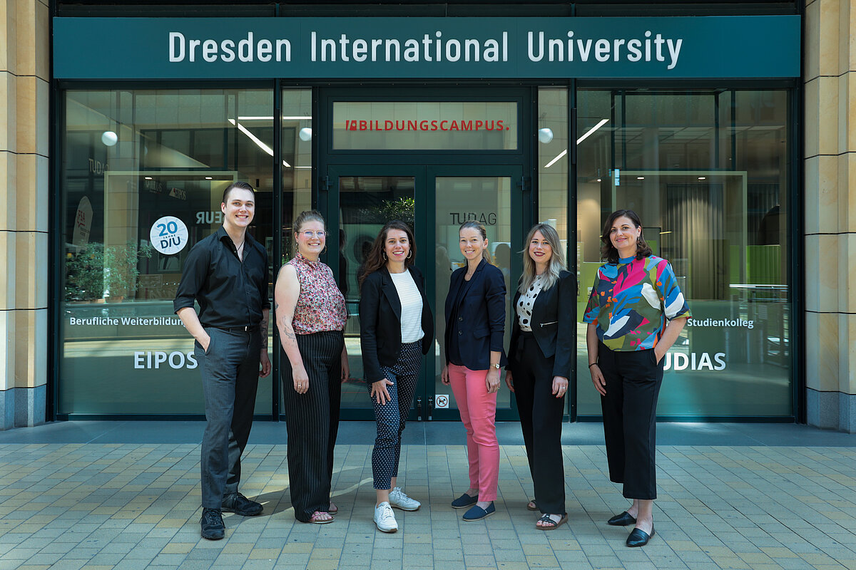 Six people standing in a row outside a building with a sign reading 'Dresden International University'