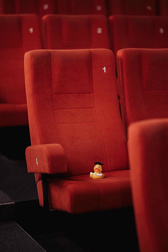 Red theater seat numbered 1 with a small rubber duck wearing a black graduation cap on it.