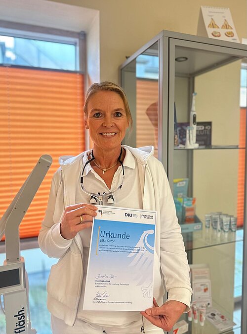 Woman in white jacket holding a certificate standing in a room with medical or dental equipment and orange window blinds