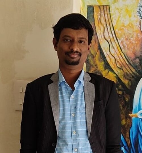 Man with short dark hair and beard wearing a black blazer over a blue checkered shirt standing indoors with a colorful painting in the background
