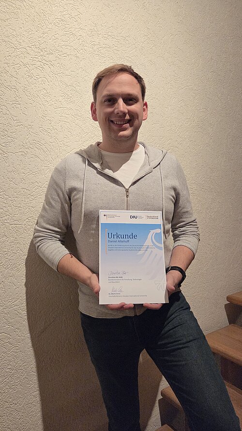 Smiling man in a light gray zip-up hoodie and dark pants holding a certificate standing next to wooden stairs against a textured wall