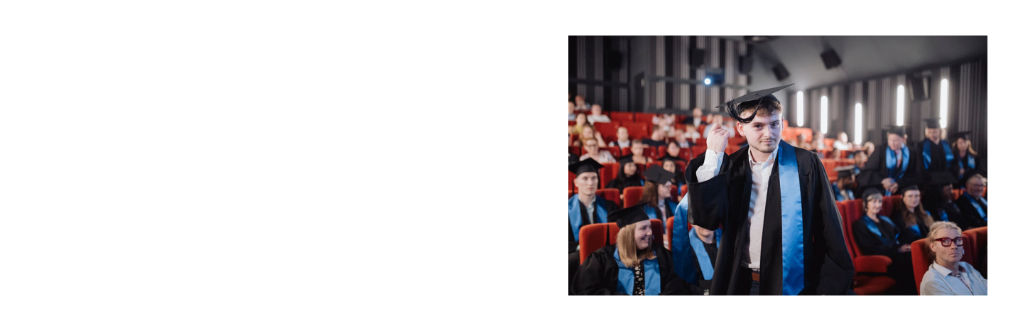 Person in graduation gown and cap standing in an auditorium with seated audience members.
