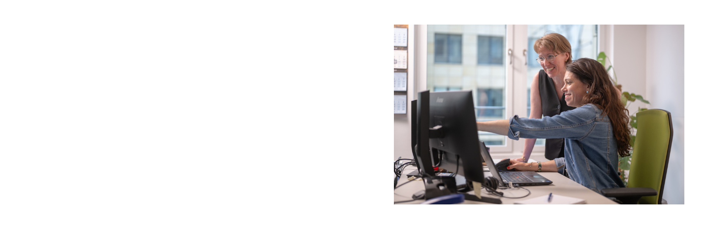 Two women at a desk with a computer one pointing at the screen while the other watches.