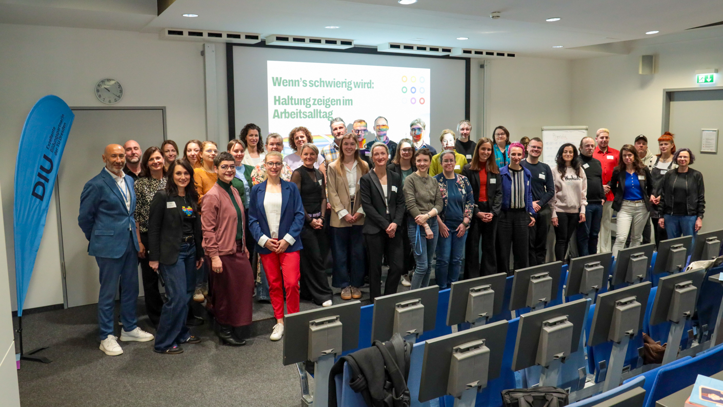 Gruppe von Menschen steht in einem Konferenzraum vor einer Leinwand mit Präsentation und blauen Stuhlreihen im Vordergrund.