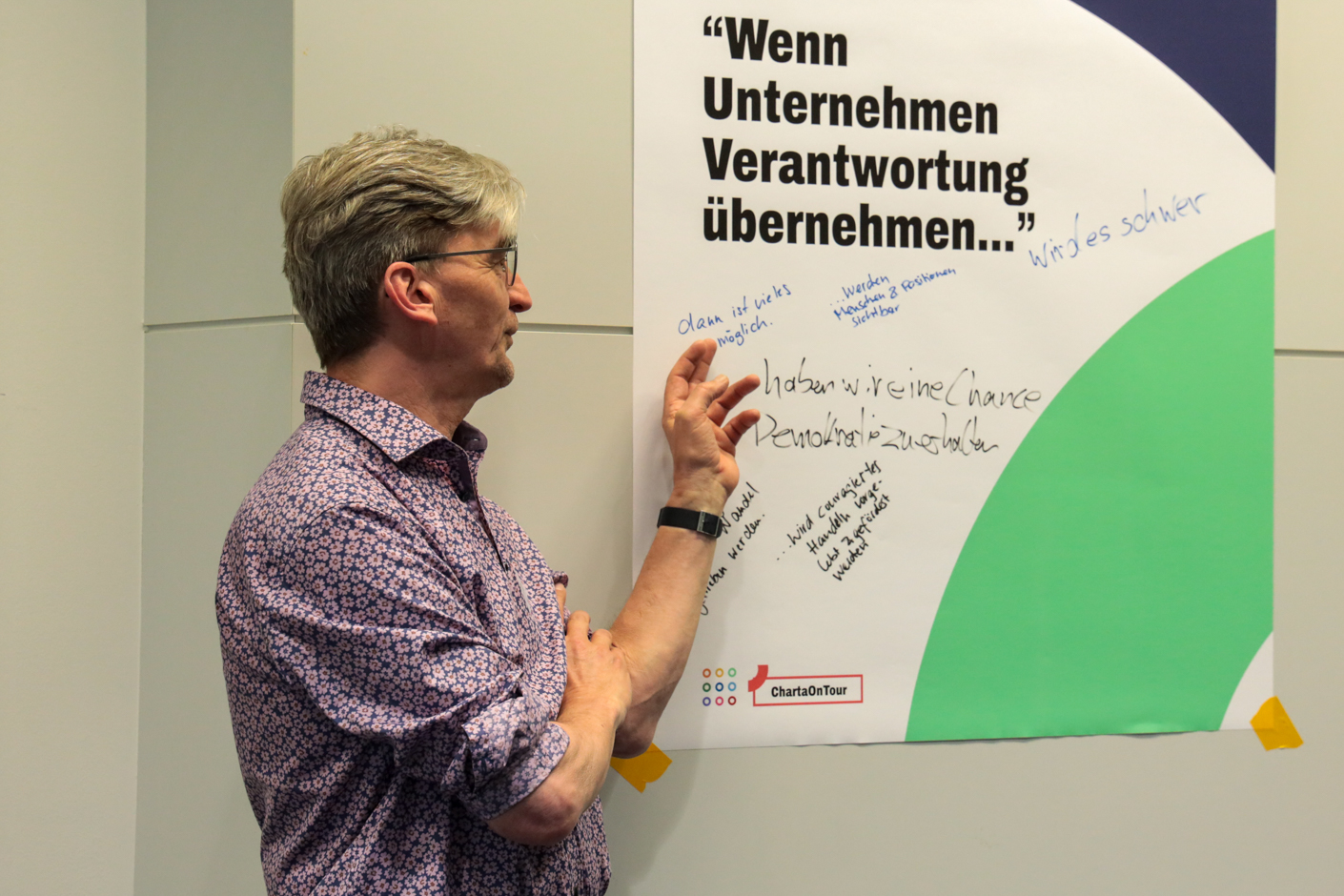 Person mit kurz geschnittenem Haar steht vor einer Wand und schreibt auf ein Plakat mit dem Text 'Wenn Unternehmen Verantwortung übernehmen'.