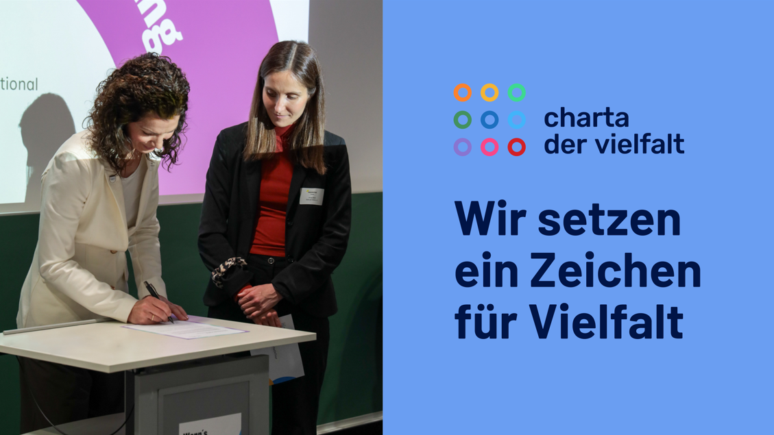 Zwei Frauen stehen an einem Tisch, eine unterschreibt ein Dokument, daneben das Logo von 'charta der vielfalt' und der Text 'Wir setzen ein Zeichen für Vielfalt'.